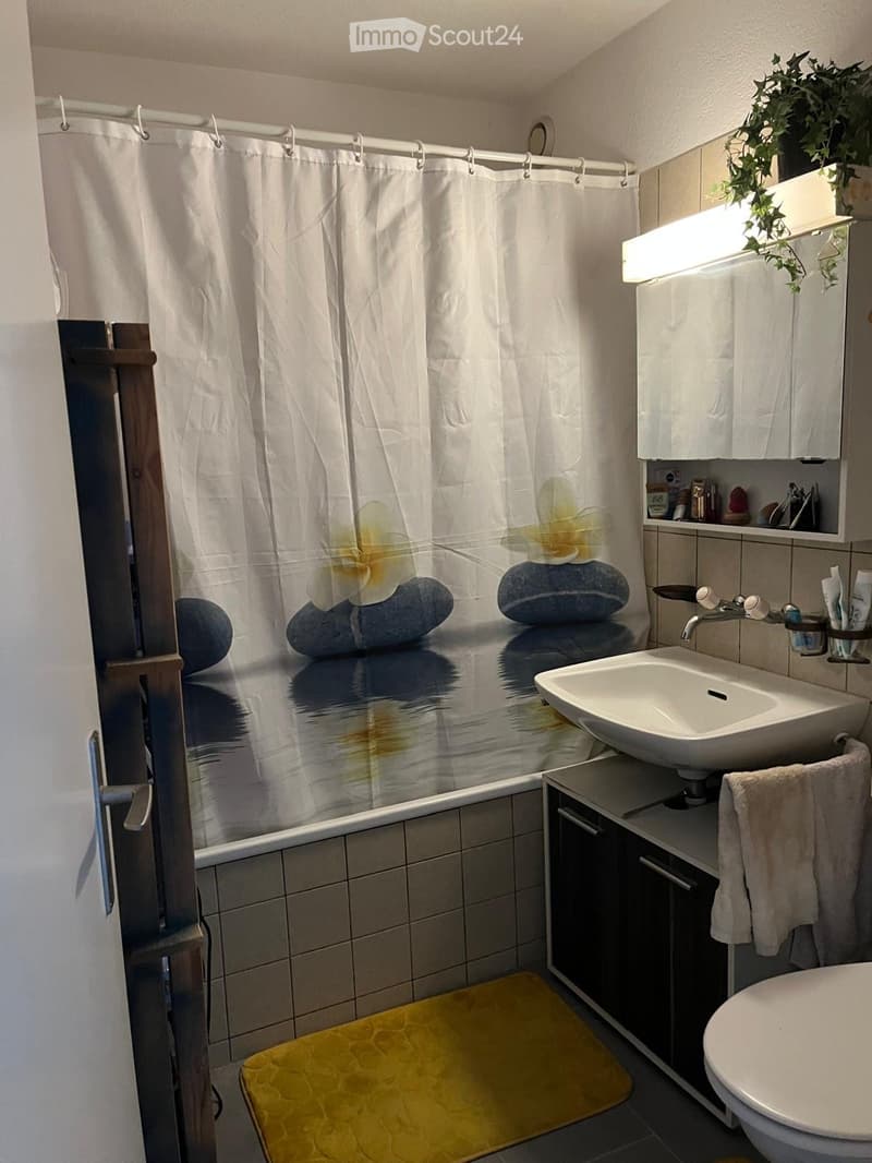 Modern bathroom with a bathtub, sink, mirror, and toilet. Features a white shower curtain with floral design, a built-in cabinet, and a yellow bathmat.
