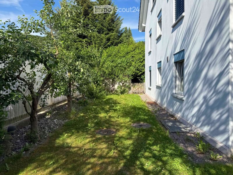 Garden area with grass, small trees, and a pathway leading to a white house with multiple windows.