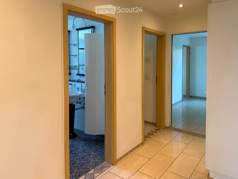 A clean, well-lit hallway with tiled flooring and wooden door frames. Visible rooms include a bathroom with a sink and a shower stall, and an adjacent empty room with tiled flooring.