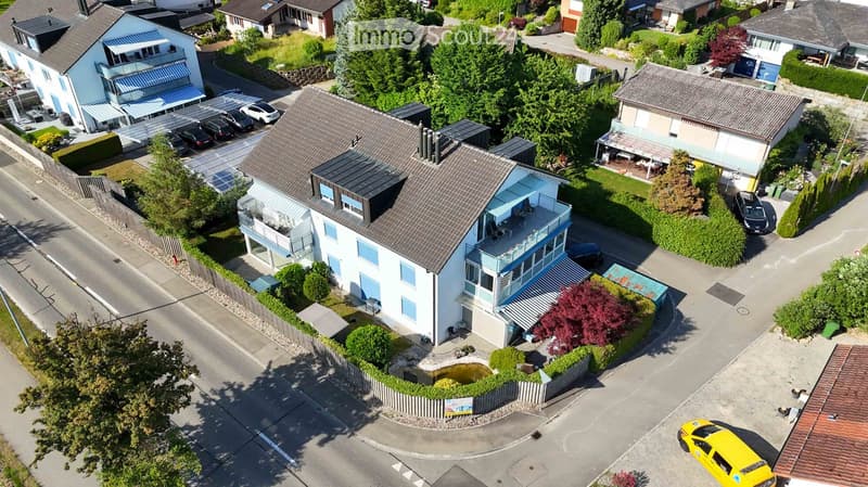 Aerial view of a modern two-story house with a balcony, surrounded by greenery and neighboring houses.