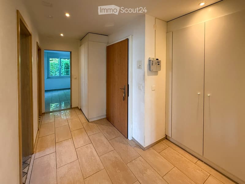 Clean hallway with tiled flooring, white walls, built-in storage cabinets, and recessed lighting. A wooden door leads to another room with a window showing greenery outside.