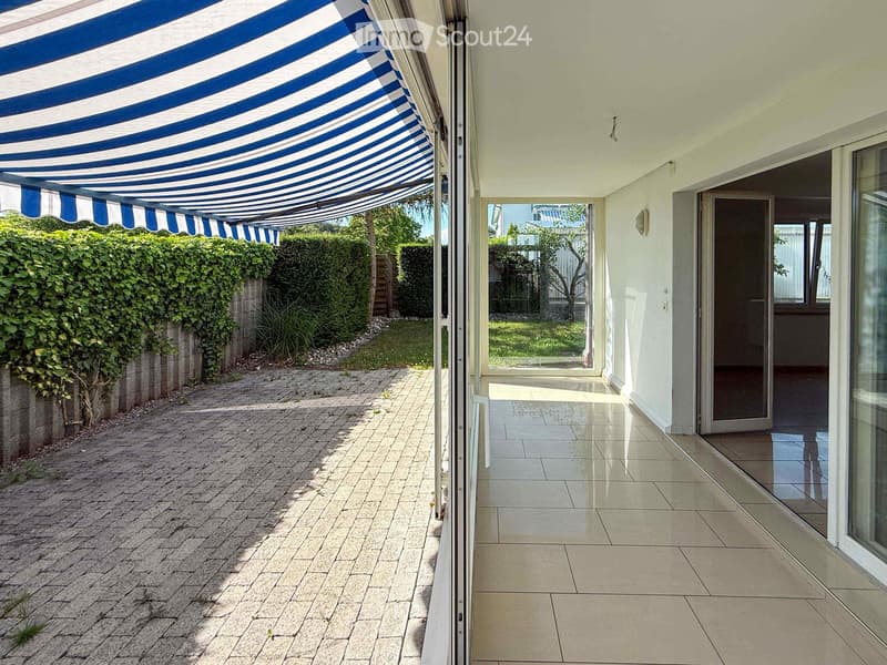 Balcony with a blue and white striped awning, tiled floor, glass doors leading to the interior, and a view of a garden with hedges and a paved pathway.