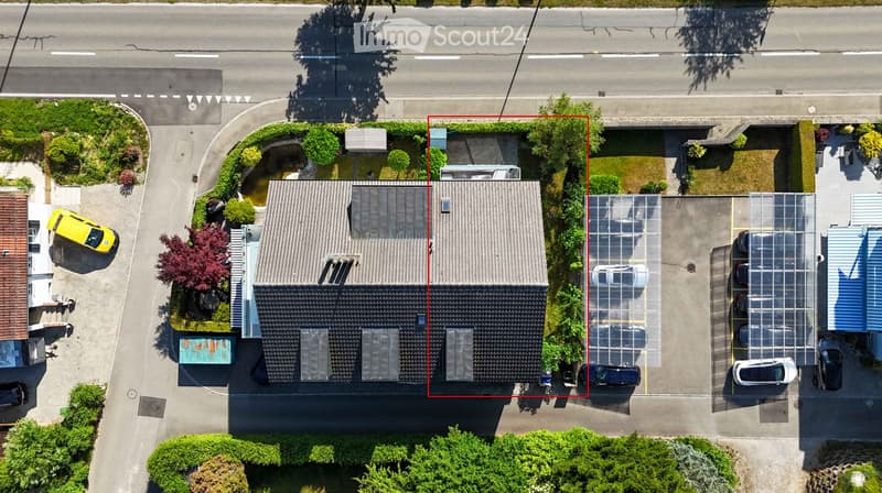 Aerial view of a two-story house with a grey roof, surrounded by greenery and a parking area with several cars.