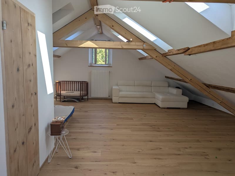 Bright attic room with wooden beams, skylights, and wooden flooring. Features a cream-colored sectional sofa, a wooden baby crib, a small glass table with a stool, and a radiator.