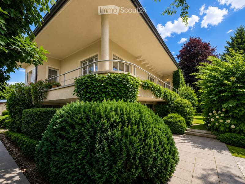 Two-story house with beige exterior, large windows, and a balcony covered in greenery. The garden features neatly trimmed hedges and a paved pathway.