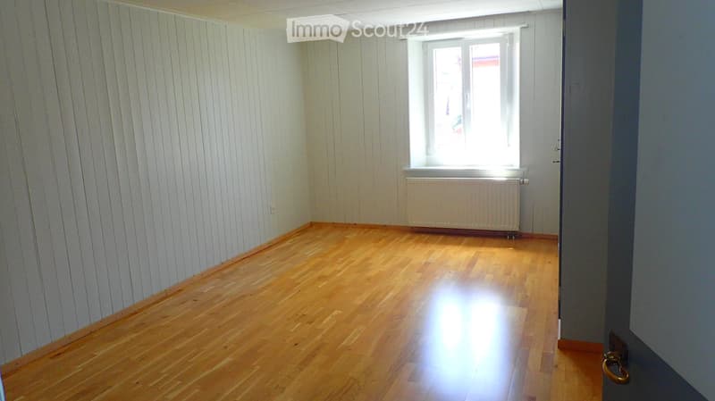 Empty room with wooden flooring, white paneled walls, a window with white frame, and a radiator below the window.