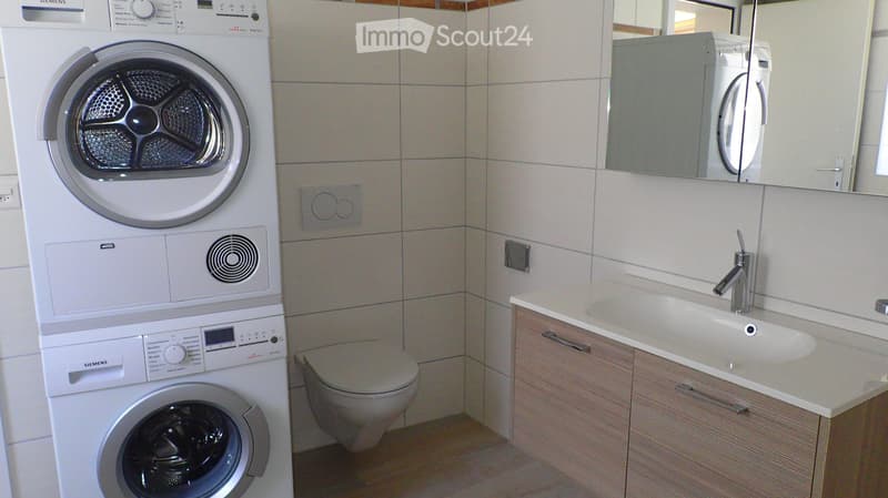 Modern bathroom with white tiled walls, a stacked washing machine and dryer, a toilet, a sink with a modern faucet, and a mirrored cabinet.