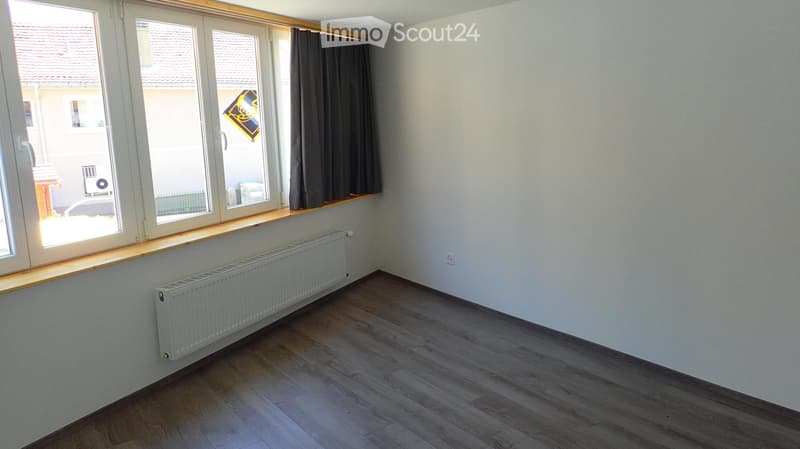 Empty room with wooden flooring, white walls, large windows with dark curtains, and a radiator under the windows.