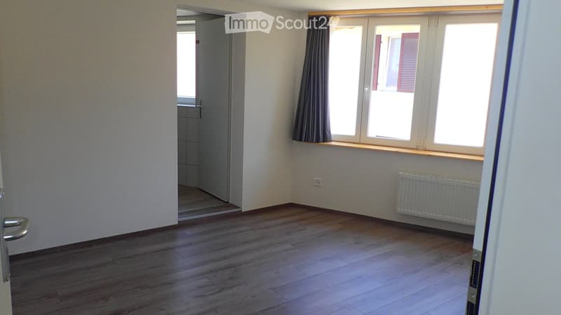 Empty room with wooden flooring, white walls, large windows with curtains, radiator, and an open doorway leading to another room.