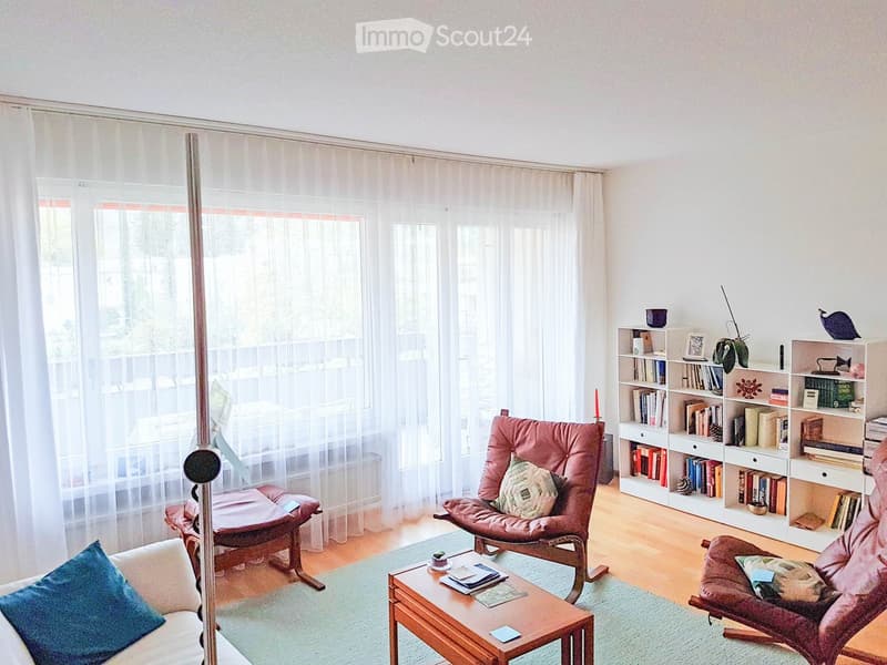 white walls, wooden floor, white curtains, large windows, balcony, armchairs, coffee table, bookshelves, books, plants
