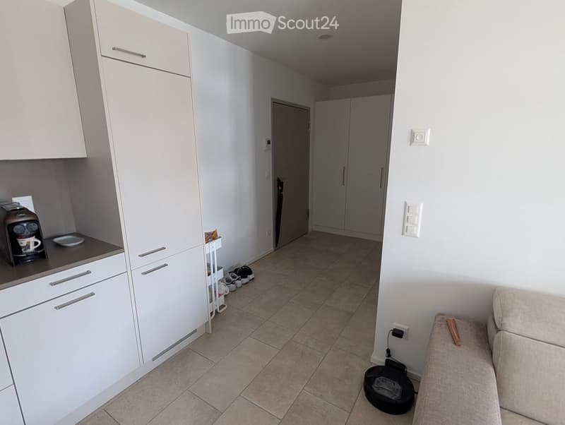 A modern, open-plan living and kitchen area with white cabinets, a coffee maker on the counter, a beige sofa, and several pairs of shoes near the entrance.