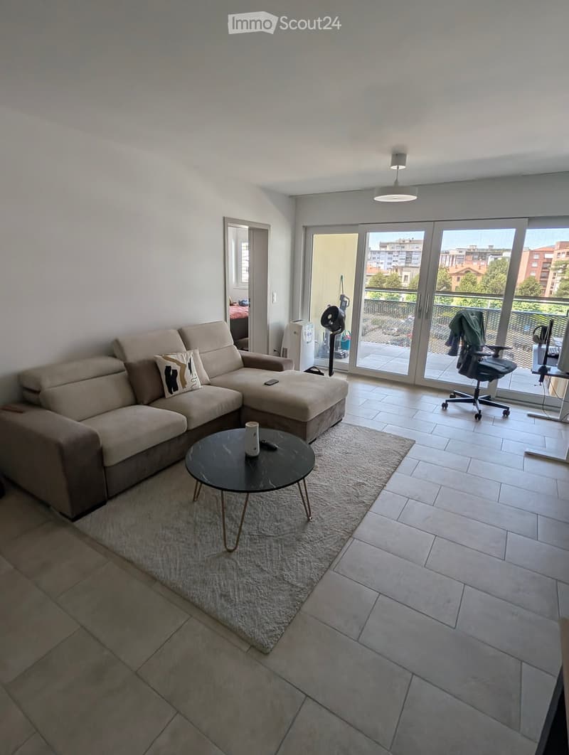 Modern living room with a beige sectional sofa, coffee table, and a small round table. The room features large sliding glass doors leading to a balcony with a view of the city.