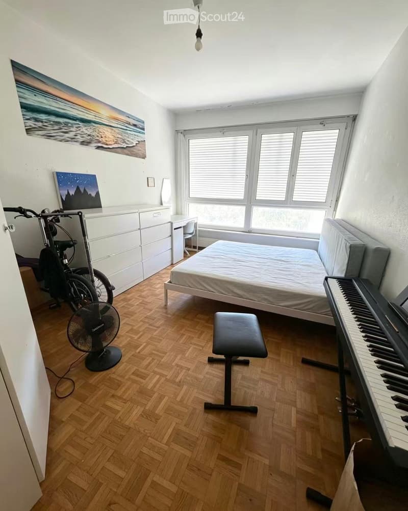 A furnished bedroom with a bed, a piano, a fan, a bicycle, and a desk area with a chair and cabinet.