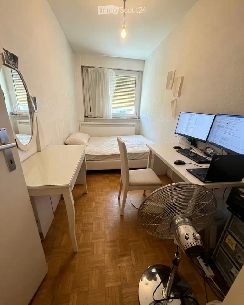 A small, well-lit bedroom with a bed, desk, chair, laptop, fan, and mirror. The room features wooden flooring and large windows with blinds.