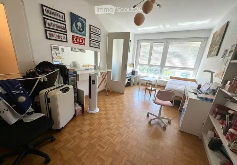 A furnished room with a wooden floor, a bed, a desk with a laptop, a chair, a suitcase, and shelves with various items. The room has large windows with blinds and several picture frames on the walls.