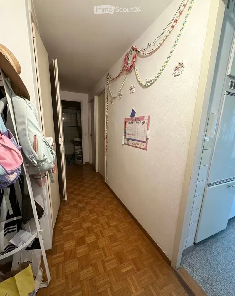 A narrow hallway with wooden flooring, light-colored walls, and a coat rack holding bags and coats. A calendar is mounted on the wall, and a small room with a sink is visible at the end.