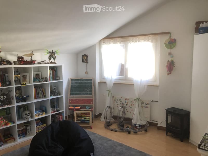A cozy children's room with a large bookshelf filled with books and toys, a beanie chair, a small table with an abacus, and a chalkboard. The room features wooden flooring, large windows with white curtains, and a colorful border along the lower wall.