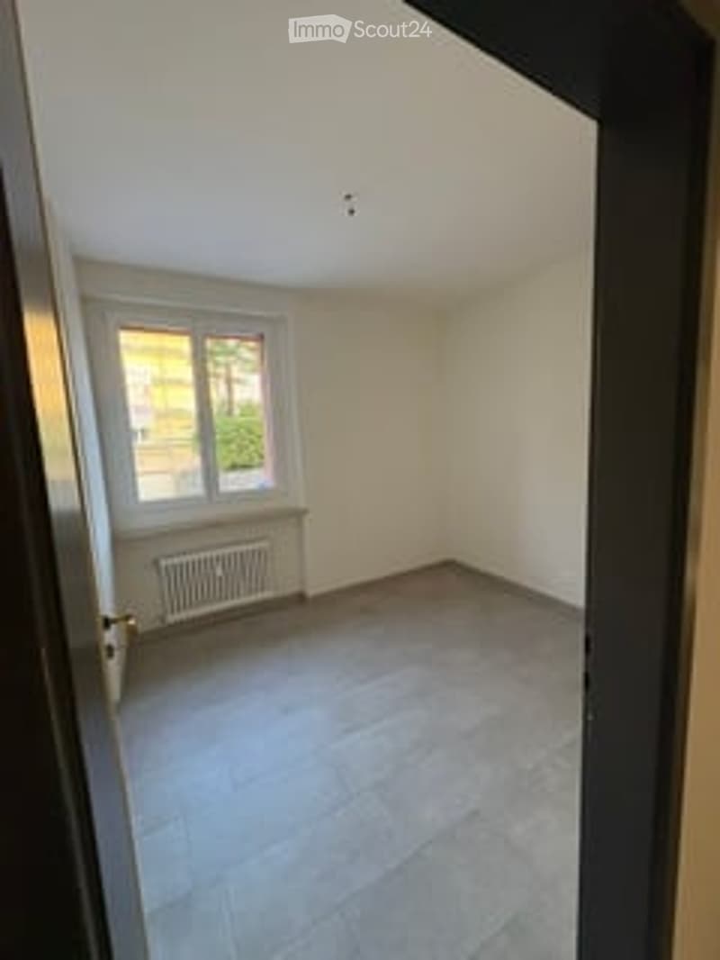 Empty room with white walls, grey tiled floor, large window, radiator, and a door leading to another space.