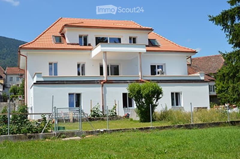 Two-story white house with red-tiled roof, multiple windows, and a balcony on the upper floor. Surrounded by a well-maintained lawn and trees, with a scenic mountain view in the background.