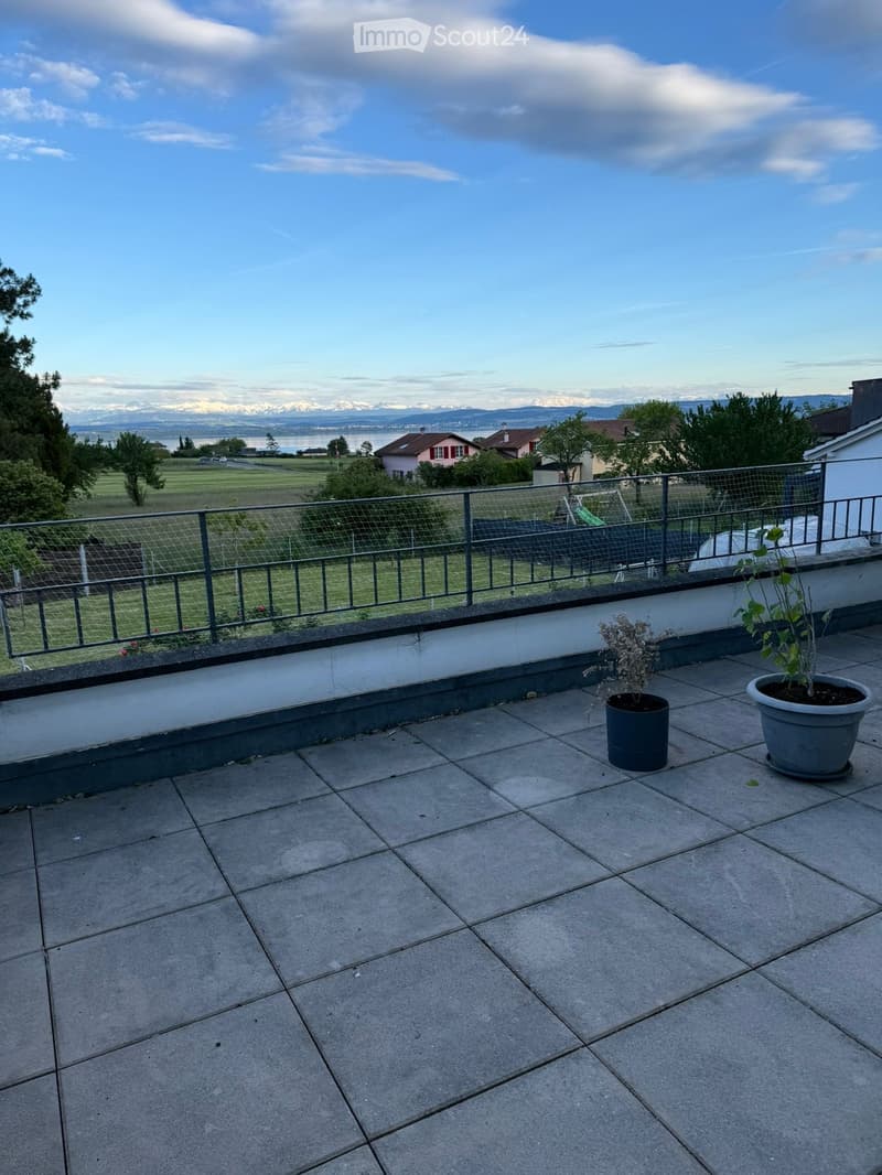 Balcony with tiled floor, metal railing, and two potted plants, overlooking a scenic landscape with mountains and a lake.