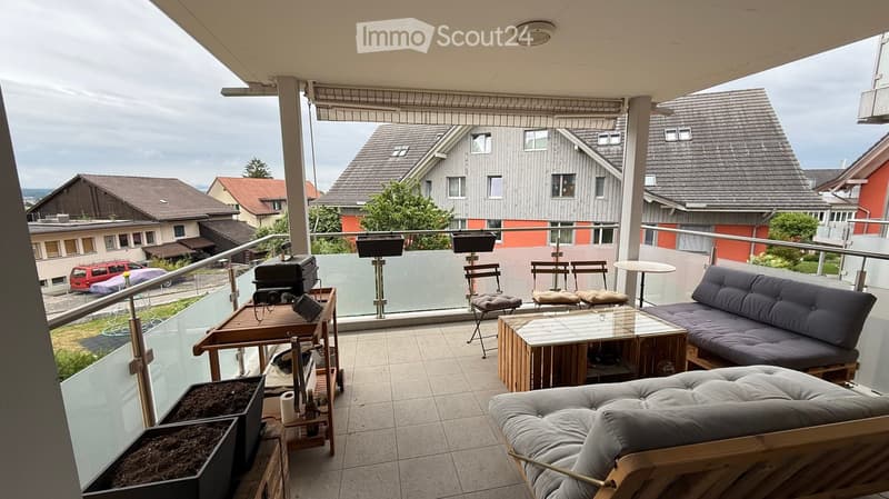 Balcony with a sofa, small table, chairs, barbecue, and flower pots, overlooking a residential area.