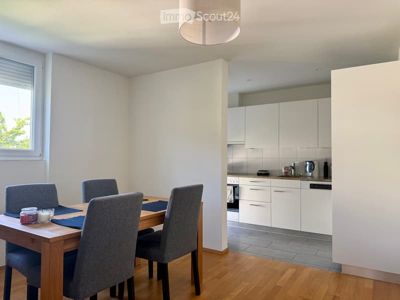 Open-plan dining and kitchen area with a wooden dining table, four grey chairs, white cabinets, and a modern kitchen setup including an oven and dishwasher.