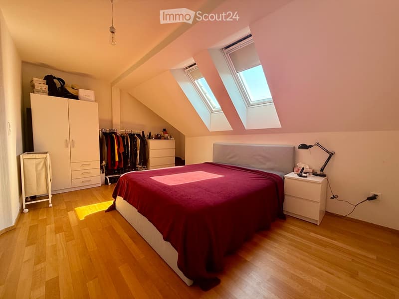 A cozy attic bedroom with wooden flooring, a large bed with a red blanket, a nightstand with a lamp, a wardrobe, a dresser, and a clothing rack with hanging clothes.