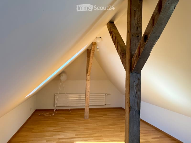 An attic room with wooden beams, wooden flooring, and large windows allowing natural light to enter. The room is empty, with no furniture or decorations.