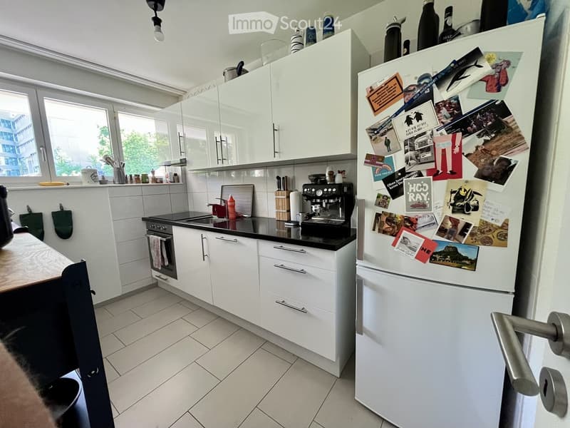Modern kitchen with white cabinets, black countertops, a refrigerator covered in magnets and photos, a coffee maker, and a small dining area with a table and chair.