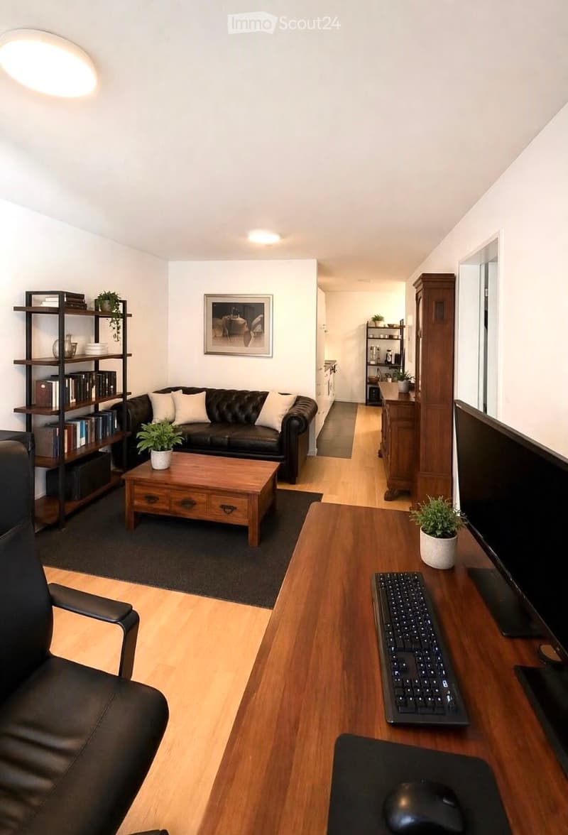 Modern living room with a black leather sofa, wooden coffee table, armchair, and a workspace area with a keyboard, mouse, and screen. Shelves with books and decorative items are visible.