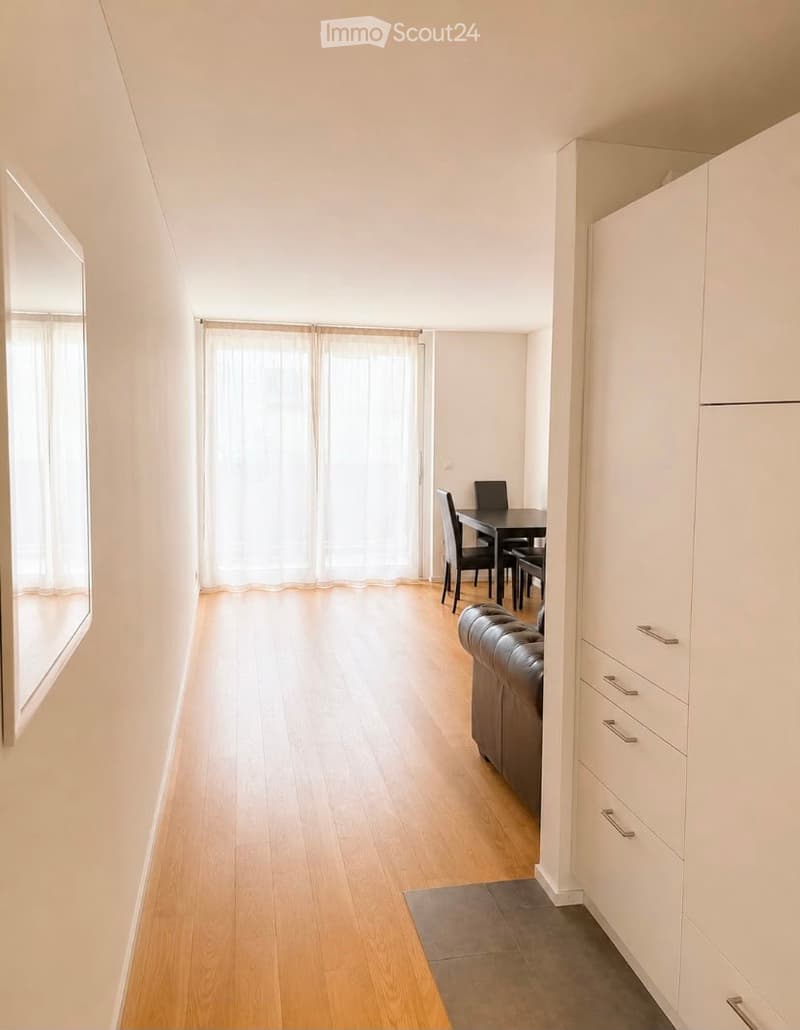 Bright, open living space with wooden flooring, large windows with white curtains, a leather sofa, and a dining area with a table and chairs.
