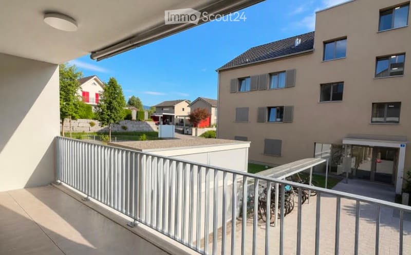 Balcony with metal railing, overlooking a residential area with modern apartment buildings, trees, and a clear blue sky.