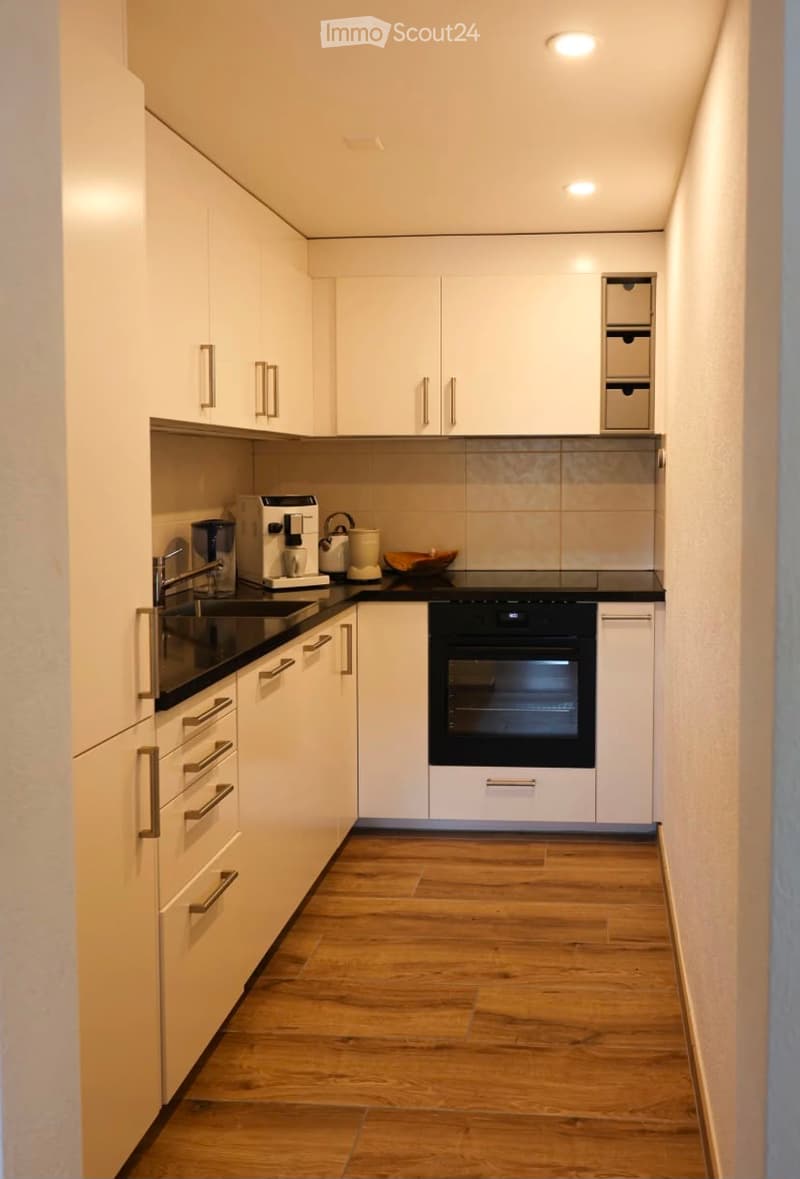 Modern kitchen with white cabinets, wooden flooring, and stainless steel appliances including a coffee maker, sink, and oven.