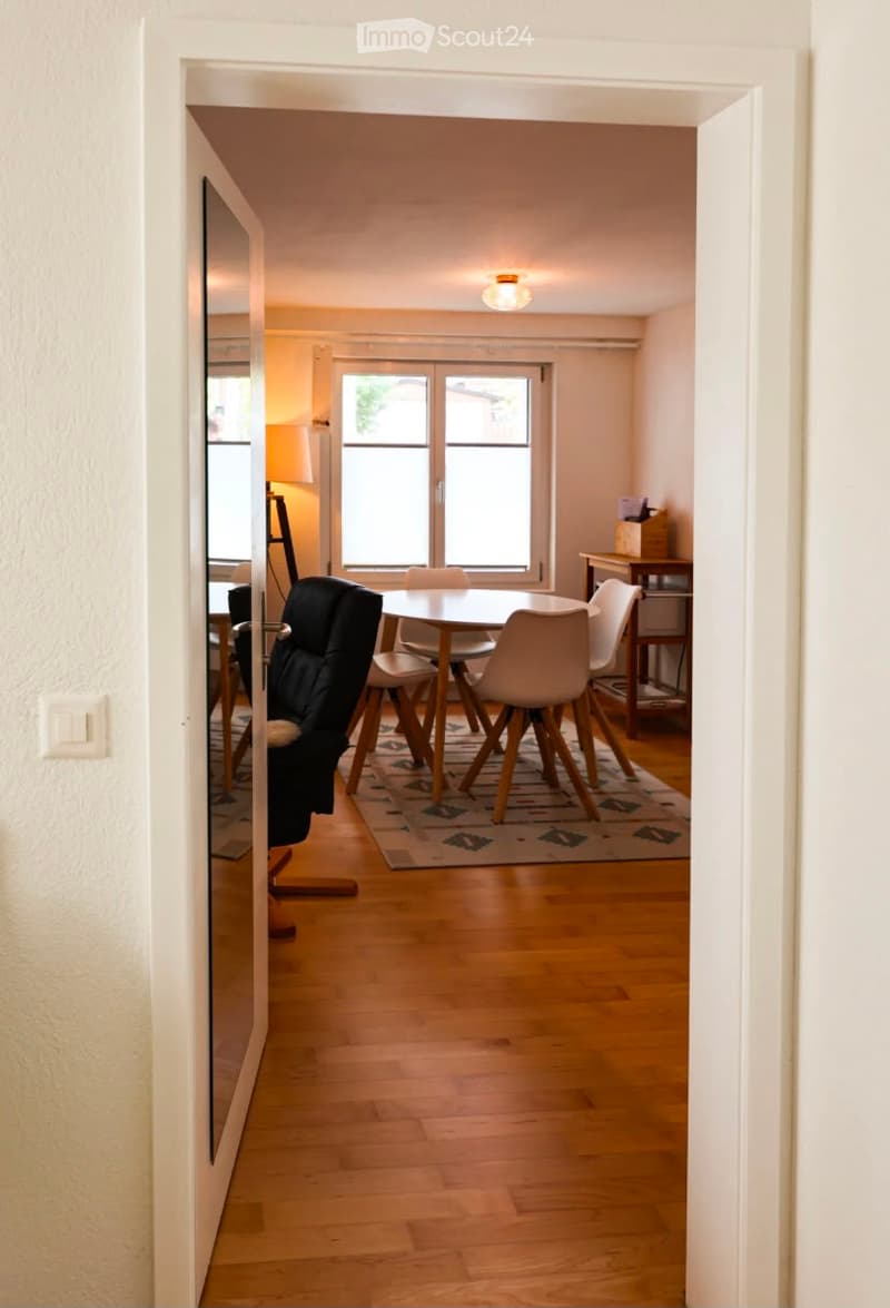 Open-plan dining area with a wooden dining table, four chairs, a lamp, and a mirror on the wall. The room features wooden flooring and large windows.