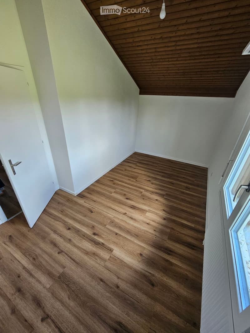 Empty room with wooden flooring, white walls, and a slanted wooden ceiling. Features a door and a window with a view.