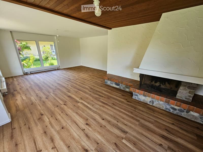 Empty room with wooden flooring, a fireplace with a stone surround, and large windows offering a view of the garden.
