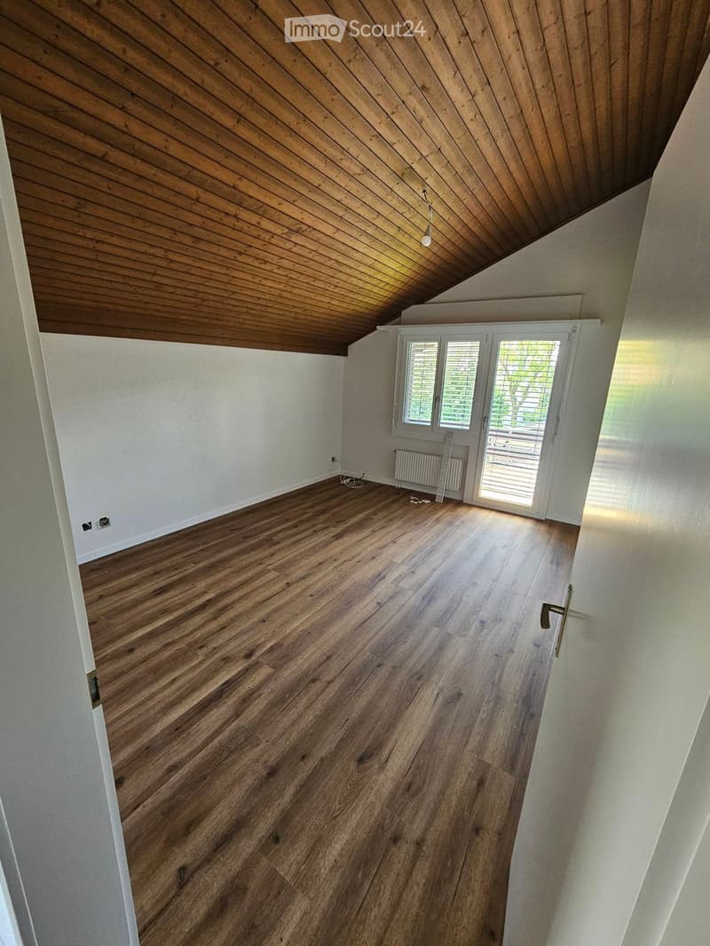 Empty room with wooden flooring, white walls, and a slanted wooden ceiling. Features a large window with blinds and a radiator under it.