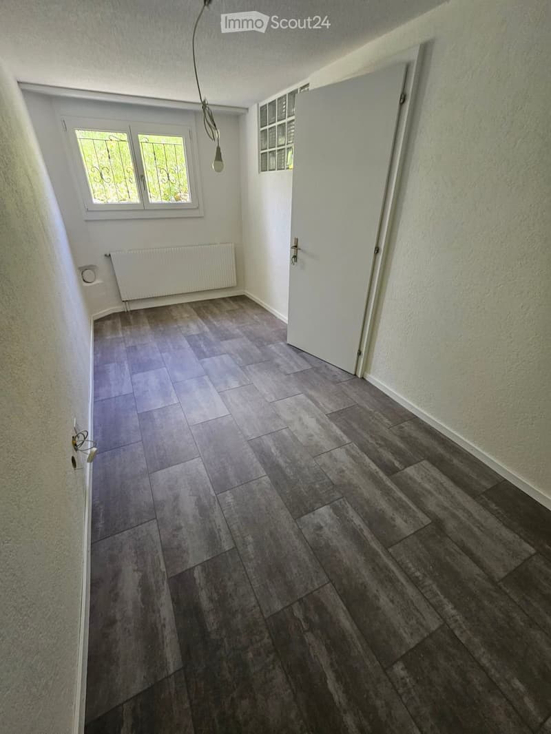 Empty hallway with tiled and wooden flooring, white walls, a window with decorative bars, a radiator, and a pendant light fixture.