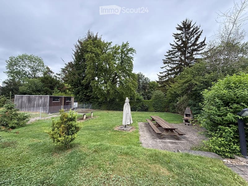 A well-maintained garden with a grassy lawn, picnic tables, a closed umbrella, a small wooden structure, and a stone fire pit surrounded by trees and bushes.