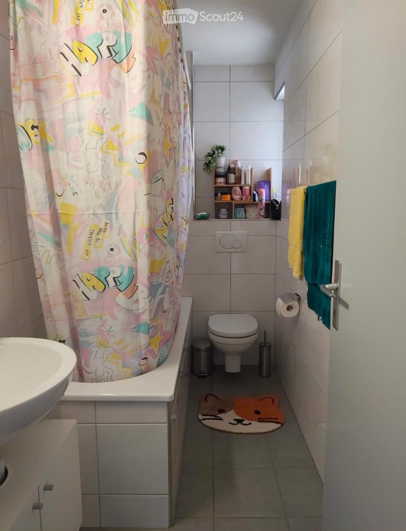 A small bathroom with a bathtub, toilet, sink, and a colorful shower curtain. A towel hangs on the wall, and a small plant sits on a shelf above the toilet.