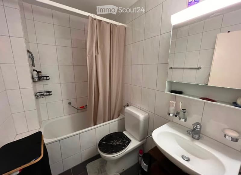 Bathroom with white tiled walls and floor, featuring a bathtub, toilet, sink, mirror, and shower curtain. Includes a shower shelf and a small trash bin.