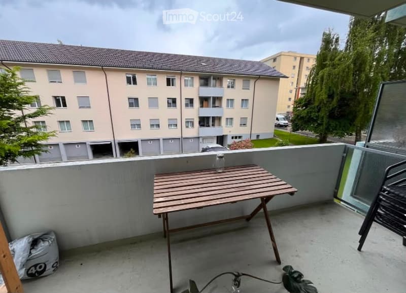 Balcony with a small wooden table and a glass jar, overlooking a residential building with multiple windows and balconies.