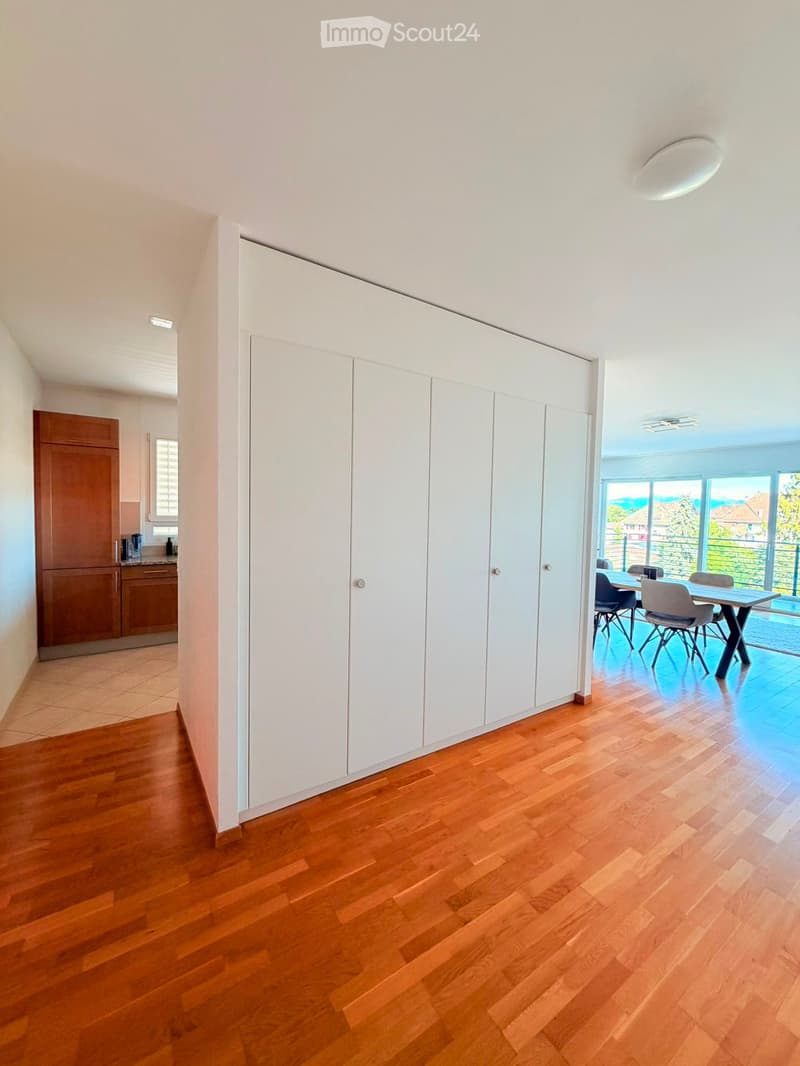 Bright hallway with wooden flooring, white built-in storage units, and a view of a dining area with a table and chairs through an open doorway.