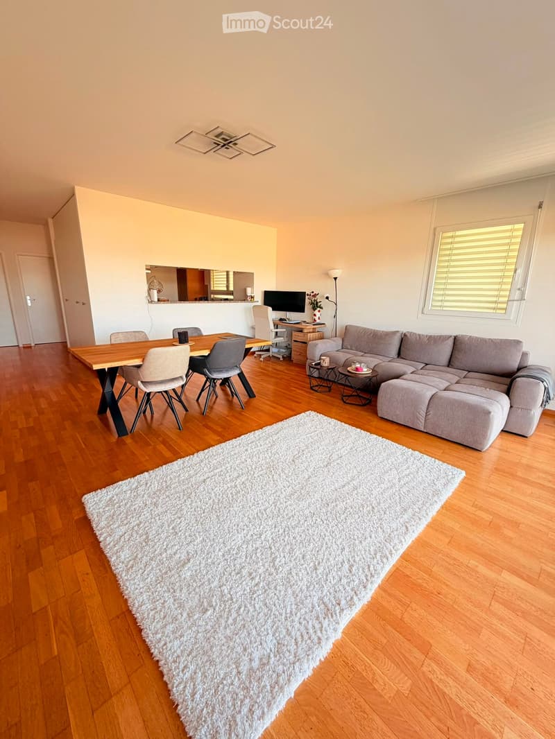 Modern living room with a large sectional sofa, coffee table, dining table with chairs, a large white shag rug, and a screen on a desk.