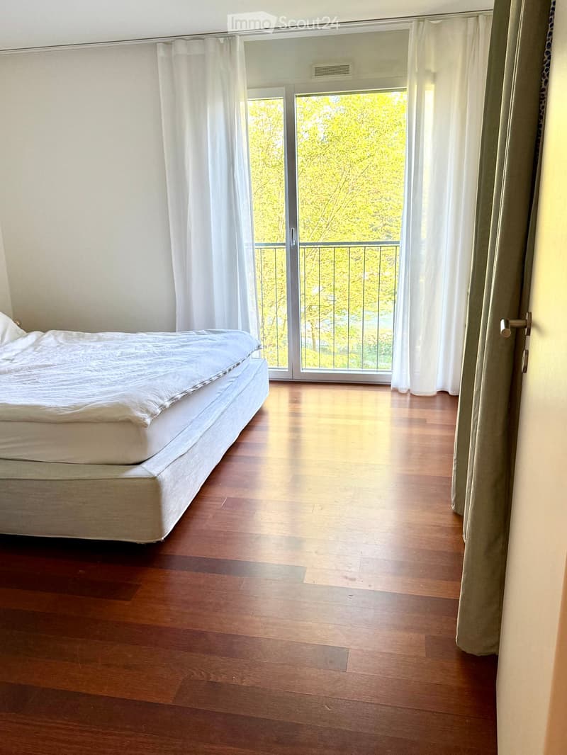 Bright bedroom with wooden flooring, a single bed, and large windows leading to a balcony with a view of trees.