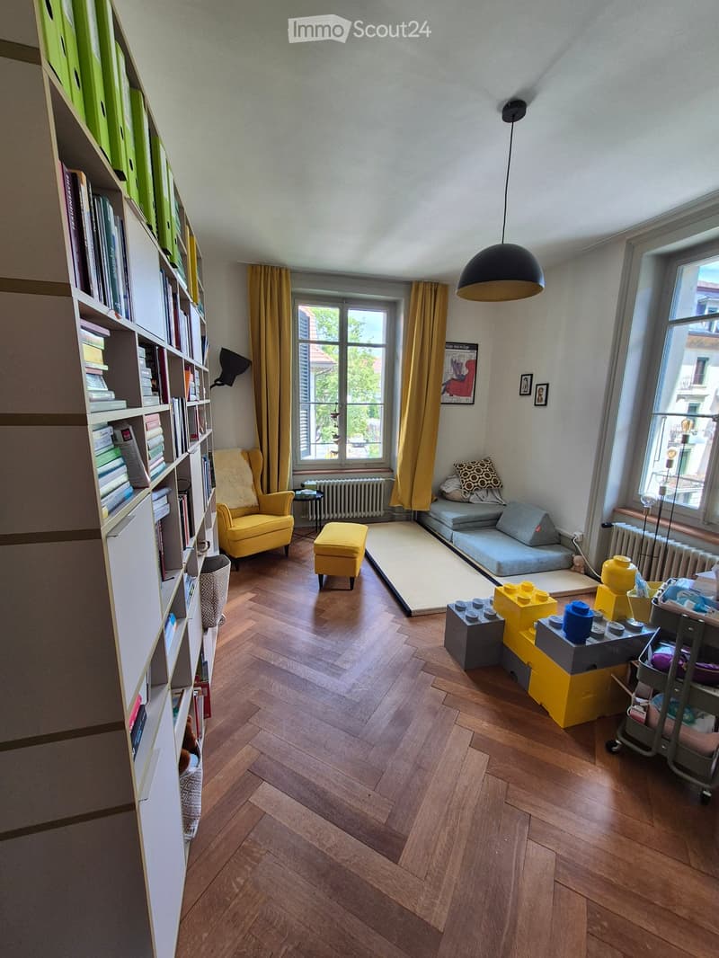 A cozy living room with wooden flooring, a yellow armchair, a small sofa, a bookshelf filled with books, and a hanging pendant light.