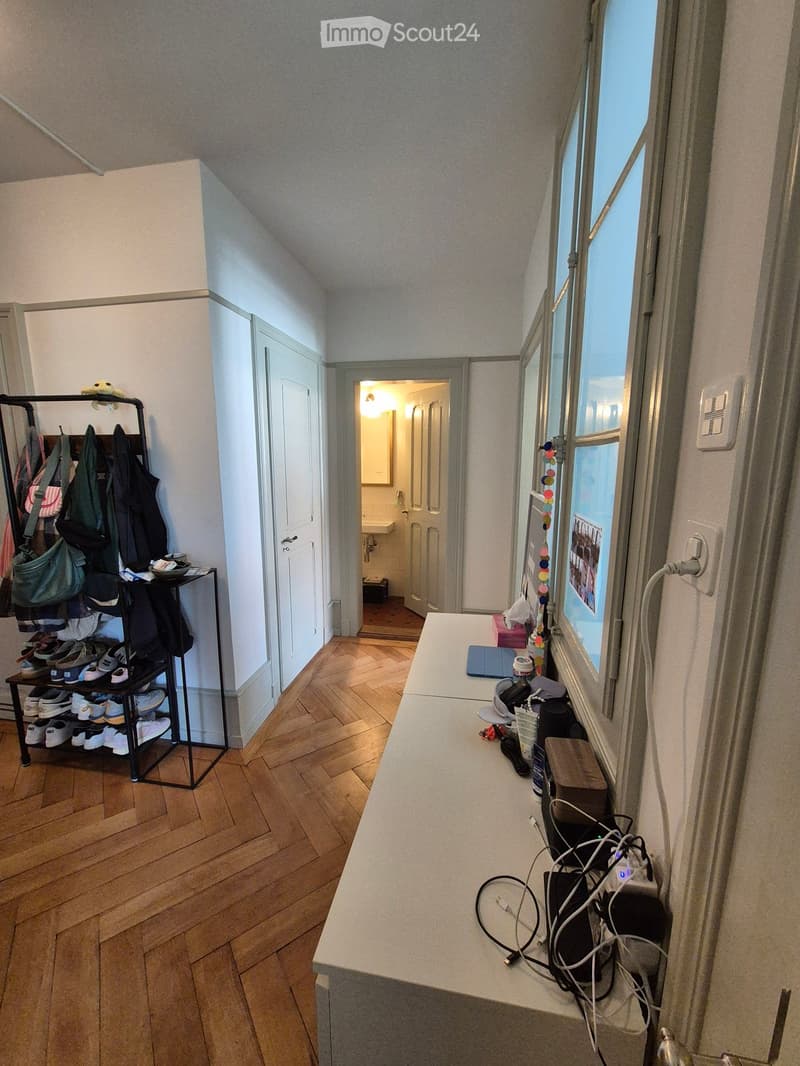 Hallway with wooden flooring, coat rack, shoe shelf, and a small table with various items, leading to a bathroom.