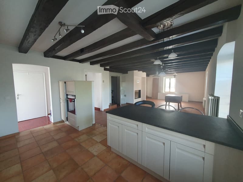 Open-plan kitchen with exposed wooden beams, tiled floor, white cabinets, black countertop, and a small dining area with a table and chairs.