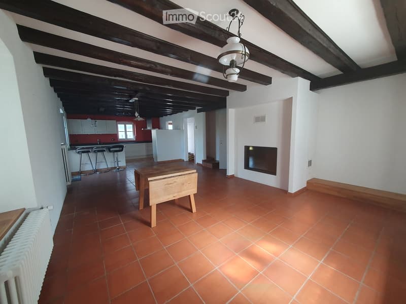 Open-plan living and dining area with terracotta tiles, wooden beams, a small wooden table, four stools, a fireplace, and a radiator. The kitchen area is visible in the background with white cabinets and a red backsplash.