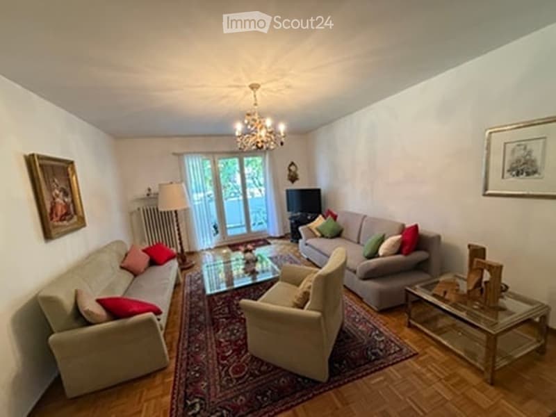 Bright living room with large windows and a balcony door, featuring a grey sofa, armchairs, coffee tables, a floor lamp, and a television. The room has a patterned rug and wooden flooring.